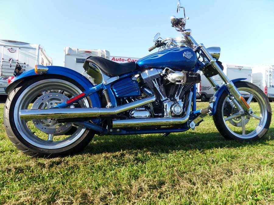 En blå motorcykel parkeret på græs med anhængere i baggrunden.