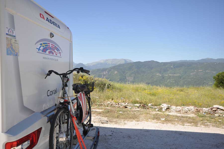 Campingvogn med cykel parkeret ved en vejkant med et bjerglandskab i baggrunden.