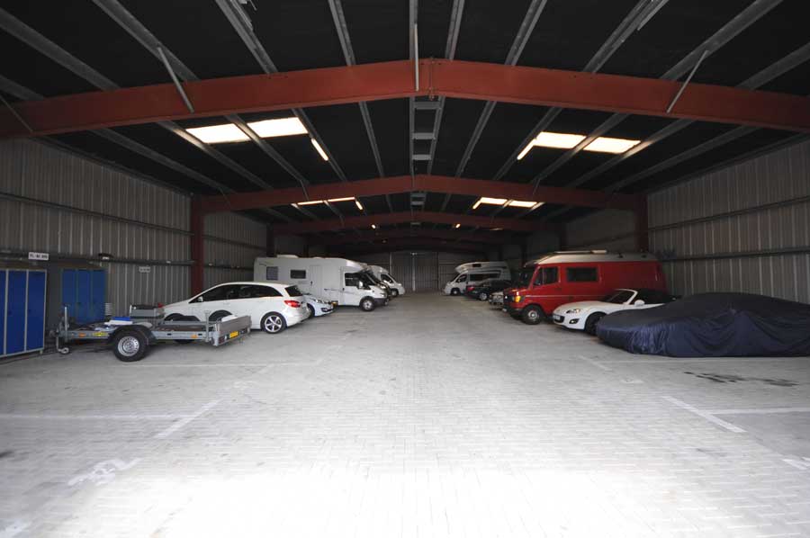 Indendørs lager med forskellige køretøjer, herunder en bil på en trailer og overdækkede biler.