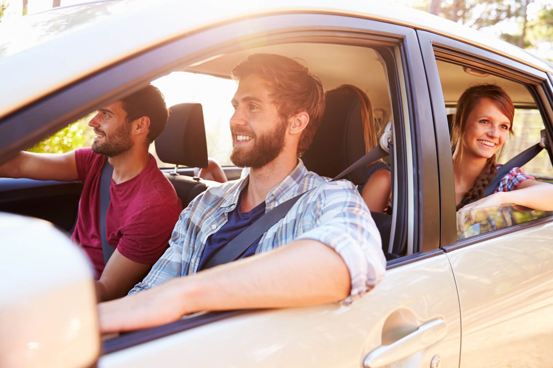Four people in a car, driver with arm out the window, passengers visible, sunny day.