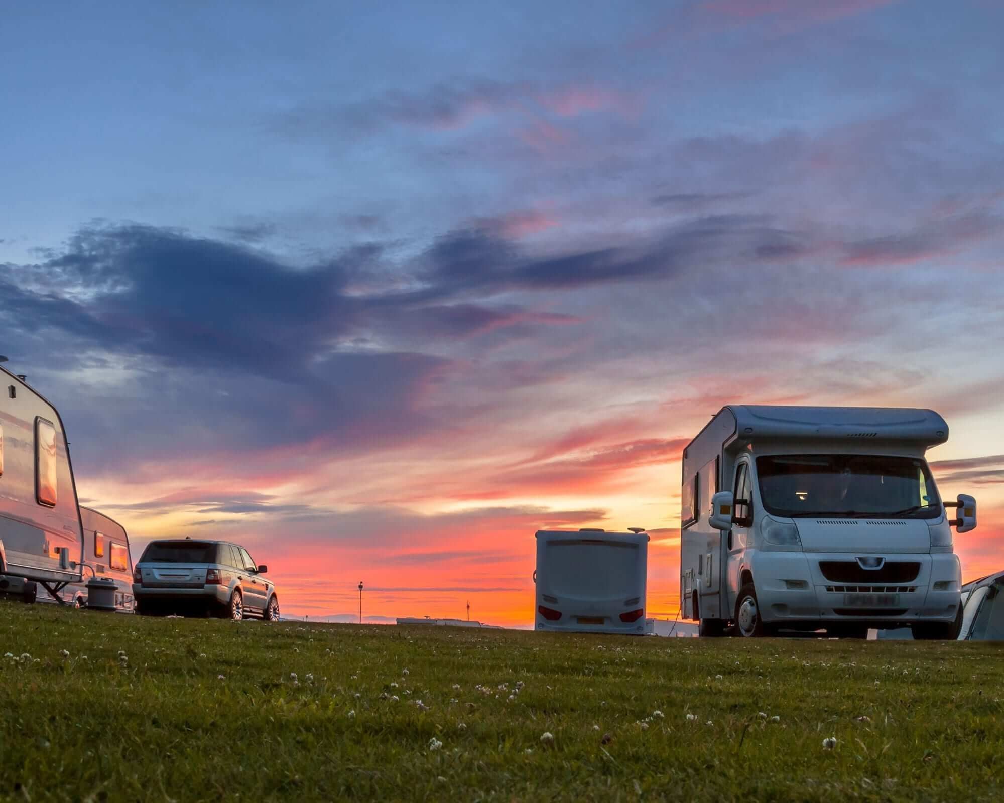 Campingvogne og en SUV parkeret på græs ved solnedgang med farverig himmel.