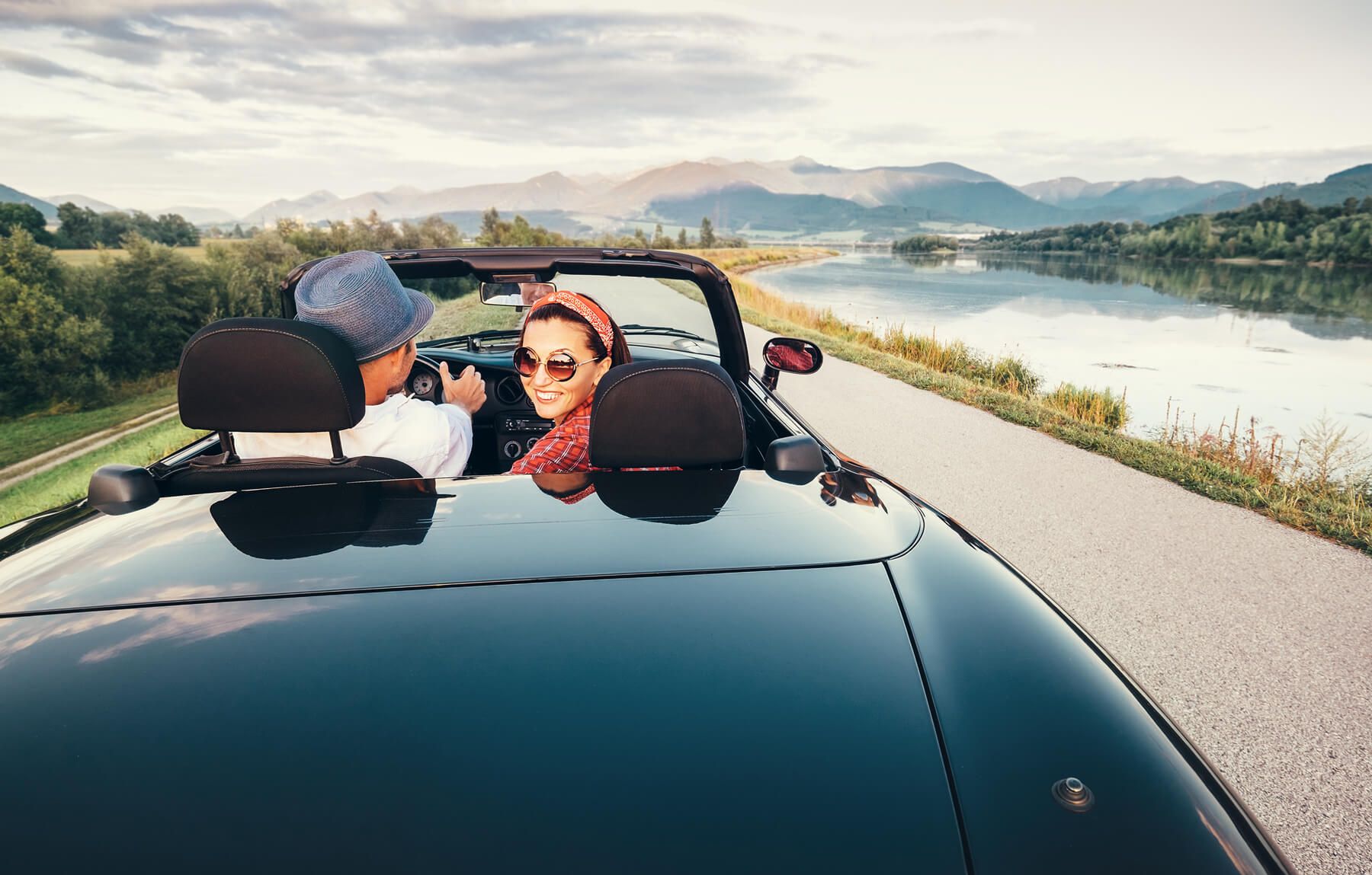 A convertible driving by a scenic lake and mountains.