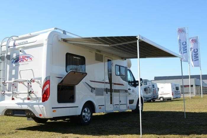 Hvid autocamper med udslået markise parkeret på græs med andre autocampere i baggrunden.